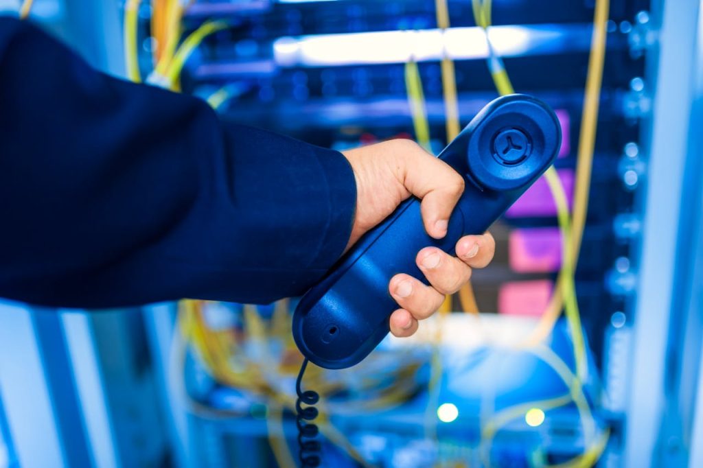 ip-phones-with-voip-technology-are-outstanding-in-international-usage-19506332 Close-up of a hand holding a telephone receiver in a server room emphasizing technology.